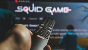 A person holds a TV remote in front of a screen showing the landing page for the Netflix (NFLX) series Squid Game.