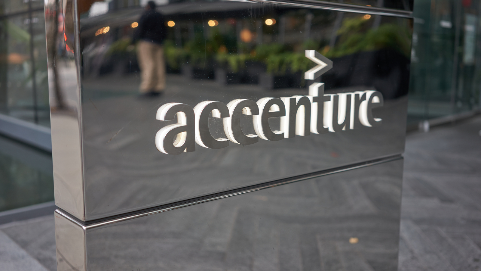 A photo of the Accenture (ACN Stock) logo in silver and white on a silver, reflective wall outside a building.