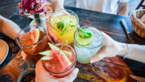 A photo of four people holding glasses with cocktails in them out in a toast, over a wooden table.