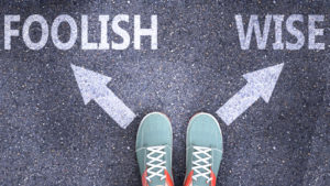 A person's shoes are seen with two arrows in front of them on the ground pointing to the words "foolish" and "wise."
