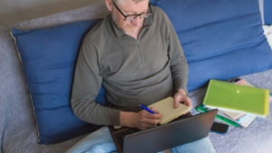 A photograph of a person sitting on a couch using a laptop and writing notes on a notepad.