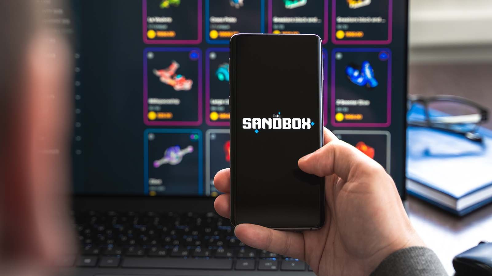 The logo for The Sandbox (SAND) on a mobile phone representing price predictions.