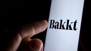 A finger touching a phone screen with the Bakkt logo on it.