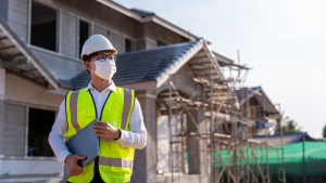 A photo of a man in a mask and neon green vest in front of a home that's under construction.