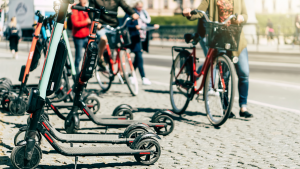 Electric scooters sit on a cobblestone walkway while people walk by with electric bikes 