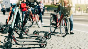 Electric scooters sit on a cobblestone walkway while people walk by with electric bikes