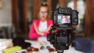 A woman in a pink jacket sits at a table inside while filming herself on camera