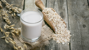 In front of a wooden background is a scoop full of oats next to a full glass of oat milk