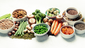 On a white table sits bowls of nuts, fruit, and seeds, while vegetables like brocolli, carrots, and mushrooms sit on the table