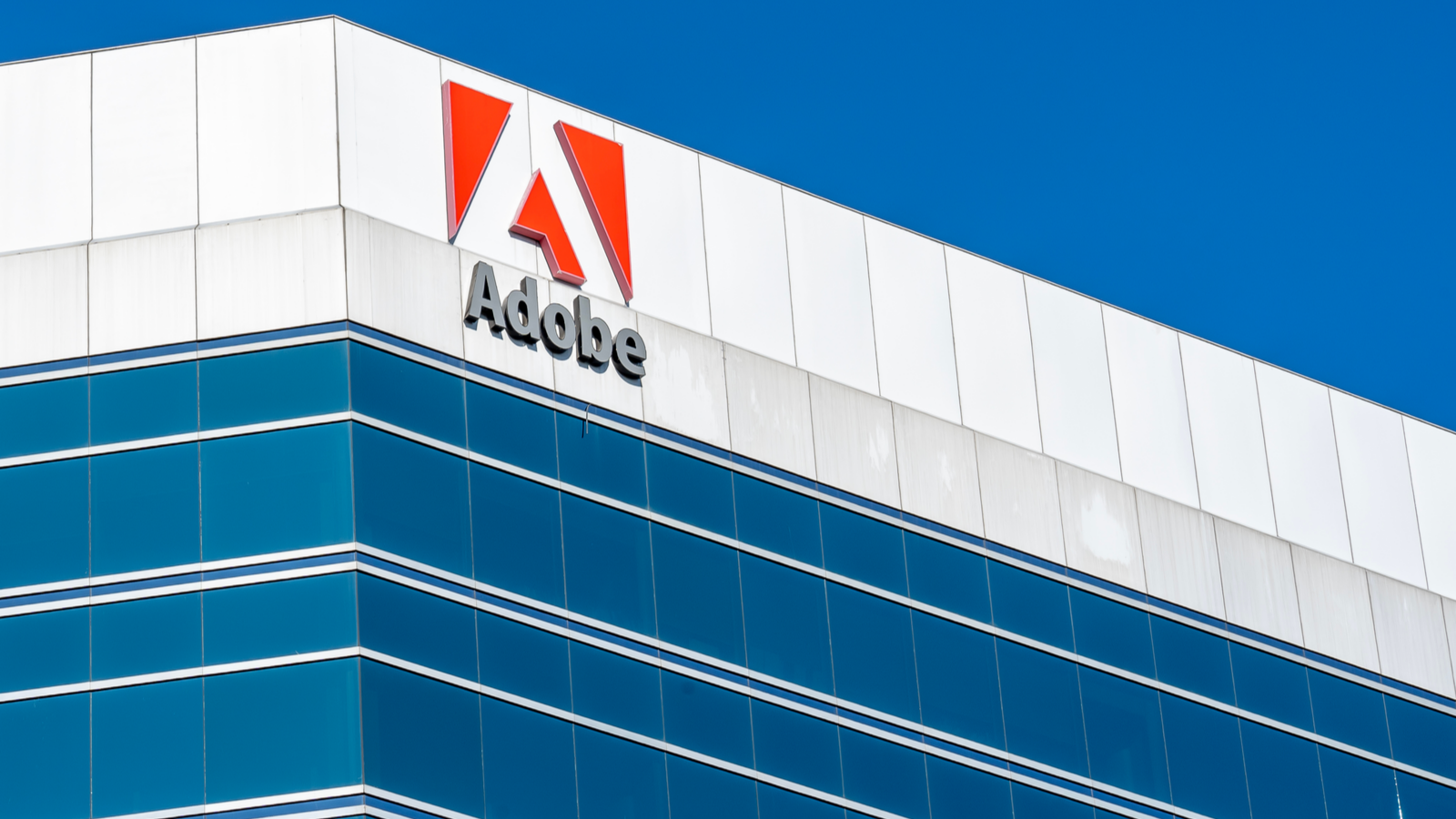ADBE Stock. A white and blue building with the Adobe logo is pictured in front of a blue sky