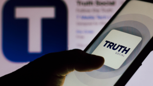 the Truth Social logo is seen displayed on a smartphone being held in front of an app store showing the app Truth Social