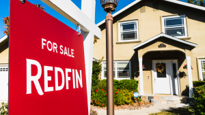 Redfin sign posted in front of a house for sale; Redfin (RDFN) is a real estate brokerage whose business model is based on sellers paying Redfin a small fee