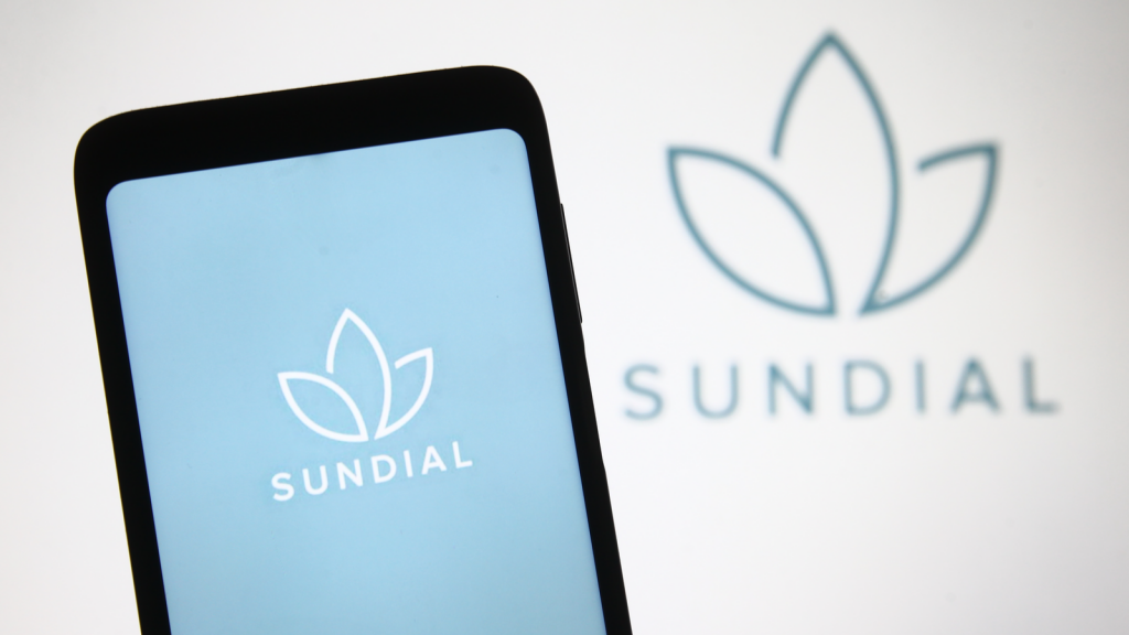 The Sundial Growers logo is on a phone screen with a light blue background in front of the sundial logo on a white background