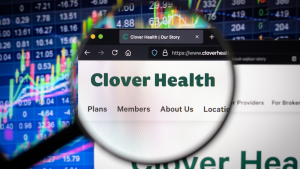 Clover Health (CLOV) company logo on a website with blurry stock market developments in the background, seen on a computer screen through a magnifying glass.