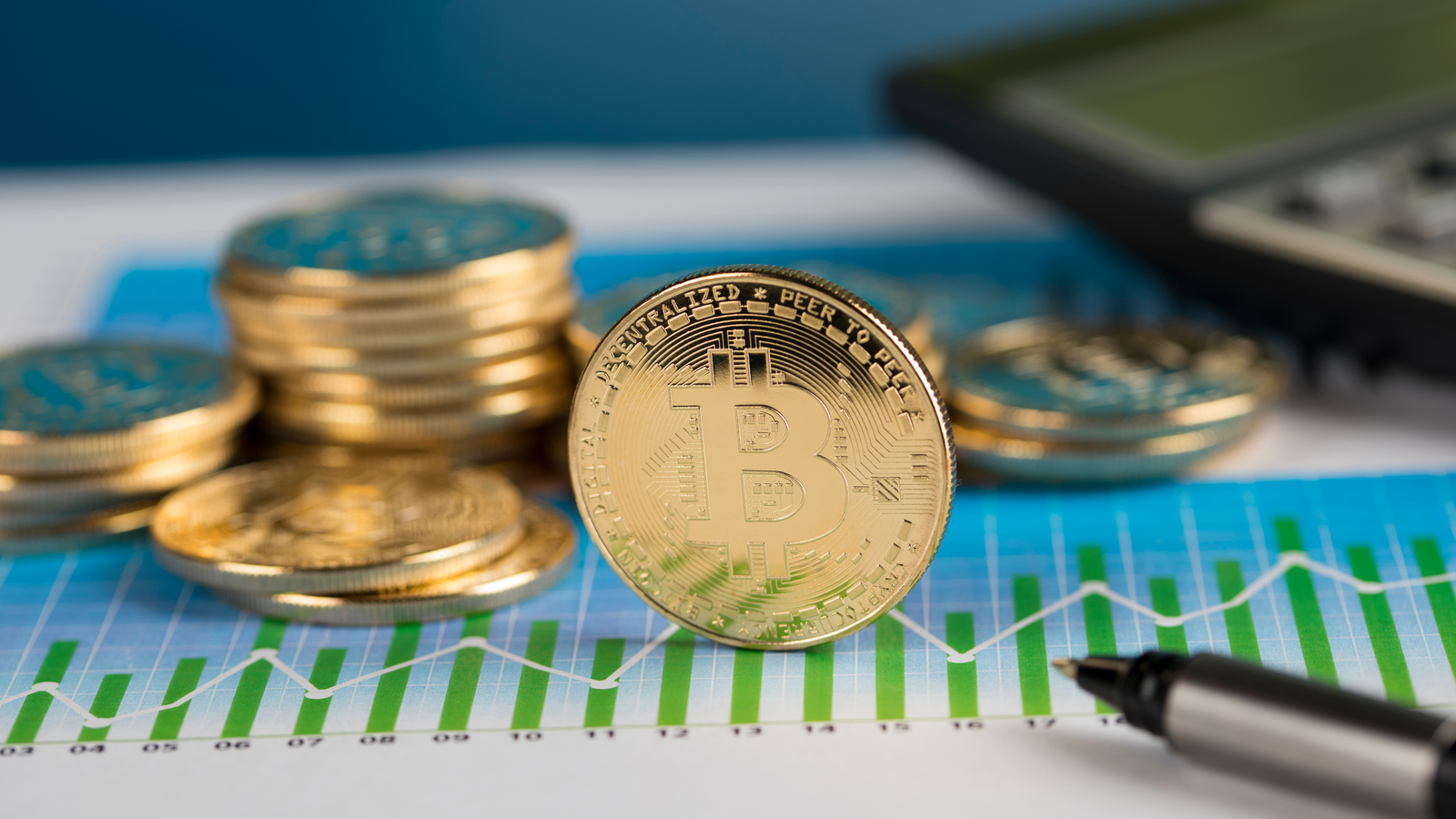 An image of a Bitcoin token standing upright on a rising graph, a calculator and pen in the background; crypto coins
