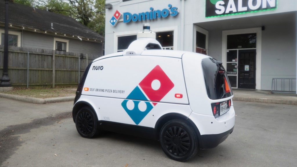 An image of a Domino's Pizza self-driving delivery car parked in front of a Domino's storefront
