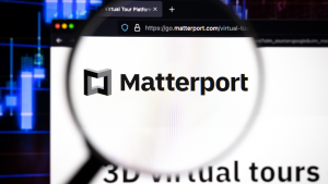 Λογότυπο εταιρείας Matterport σε έναν ιστότοπο με θολές εξελίξεις στο χρηματιστήριο στο παρασκήνιο, που φαίνεται σε μια οθόνη υπολογιστή μέσα από ένα μεγεθυντικό φακό.  Απόθεμα MTTR.