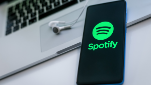 Spotify (SPOT) のロゴが表示されたスマートフォンの拡大図。 背景にラップトップとヘッドフォン。 新しいテクノロジー、ソーシャル メディア、ネットワーク、リキッド ミュージックのコンセプト。