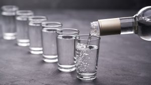 A bottle of vodka being poured into a row of shot glasses.