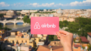 Person holding Airbnb logo over the cityscape of Rome, Italy. ABNB stock.