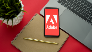 The Adobe logo on the smartphone screen is placed on the keyboard of an Apple MacBook on a red desk background.  ADBE stock.