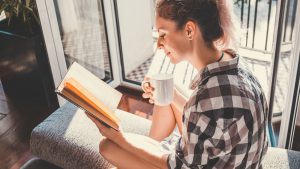 woman sitting at opened window drinking coffee and reading a book