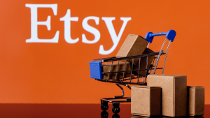 オレンジ色の背景の上に Etsy のロゴがあり、パッケージが入った小さなショッピング カートが表示されます。 ETSY株。