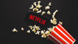 An image of a phone with the Netflix logo on the screen, lying next to a container of popcorn with popcorn splayed across