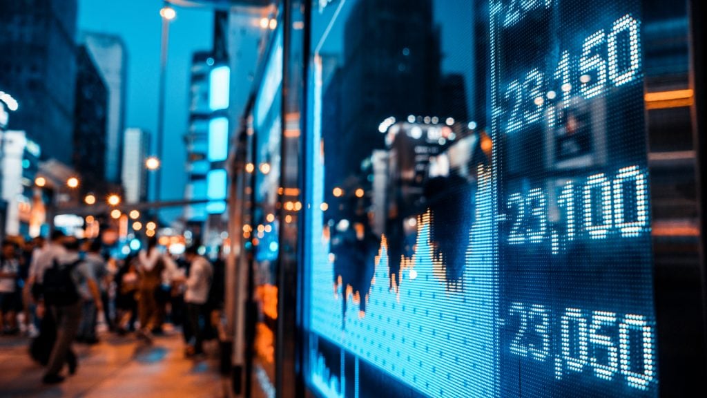 A photo of stock prices on a digital screen with a city street reflected in the glass and visible in the left side of the image; stock market prices