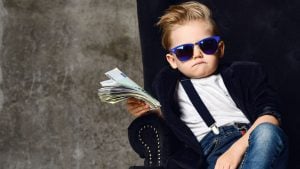 A photo of a young boy wearing sunglasses, jeans, blazer, white shirt and suspenders holding money in different denominations in one hand and sitting in a soft chair.