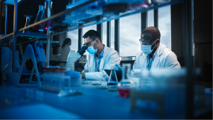 OLK Stock. Modern Medical Research Laboratory: Two Scientists Wearing Face Masks use Microscope, Analyse Sample in Petri Dish, Talk. Advanced Scientific Lab for Medicine, Biotechnology. Blue Color. KZR stock.