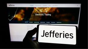 A photo of a hand holding a smartphone showing the Jefferies logo while in the background, a monitor shows the Jefferies website.