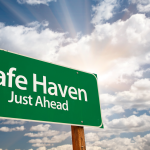 Road sign that says Safe Haven Just Ahead in front of a backdrop of blue sky and clouds. Safe haven stocks. Safe stocks.