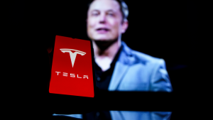The Tesla Inc. (TSLA) logo displayed on a phone in front of a blurry image of Elon Musk