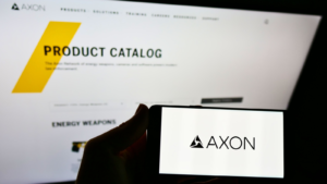 Person holding mobile phone with logo of American weapon manufacturer Axon Enterprise Inc. on screen in front of webpage. Focus on cellphone display. Unmodified photo.