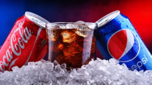 KO stock PEP stock: a can of Coca-cola and a can of Pepsi on either side of a glass of brown soda and sitting on top of a pile of ice