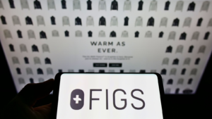 Person holding smartphone with logo of US healthcare apparel company Figs Inc. on screen in front of website. Focus on phone display. Unmodified photo.