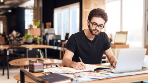 Freelancer bearded man in t-shirt taking notes at laptop sitting at desk. FLNCF stock