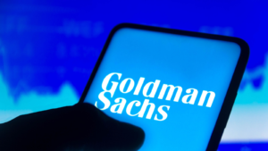 In this photo illustration, the Goldman Sachs Group (GS) logo is displayed on a smartphone screen and a stock market chart in the background