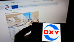 ウェブサイトの前の画面にアメリカの企業オキシデンタル・ペトロリアム・コーポレーション（OXY）のロゴが表示された携帯電話を持っている人。 電話のディスプレイに注目してください。 無修正の写真。
