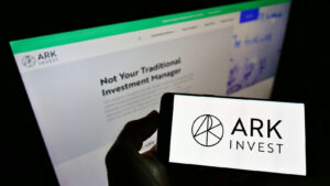 Ark Invest logo displayed on mobile phone with computer monitor showing Ark Invest website in background