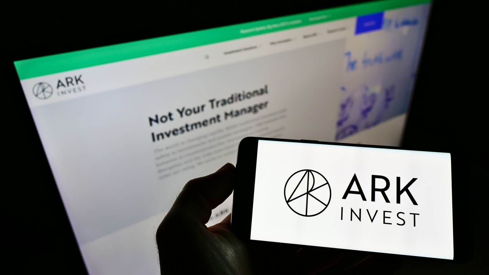 Ark Invest logo displayed on mobile phone with computer monitor showing Ark Invest website in background