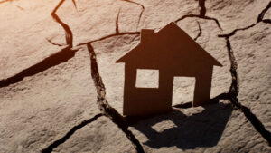 Flat cut-out image of house jammed into the crack of dry desert, symbolizing housing crisis