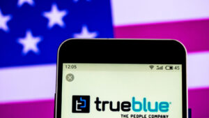 TBI stock: the TrueBlue logo on a phone in front of an american flag background