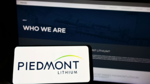 米国の鉱業会社 Piedmont Lithium Inc. (PLL) のロゴが画面に表示された携帯電話を、企業のウェブページの前に掲げている人物。携帯電話のディスプレイに焦点を合わせている。加工なしの写真。