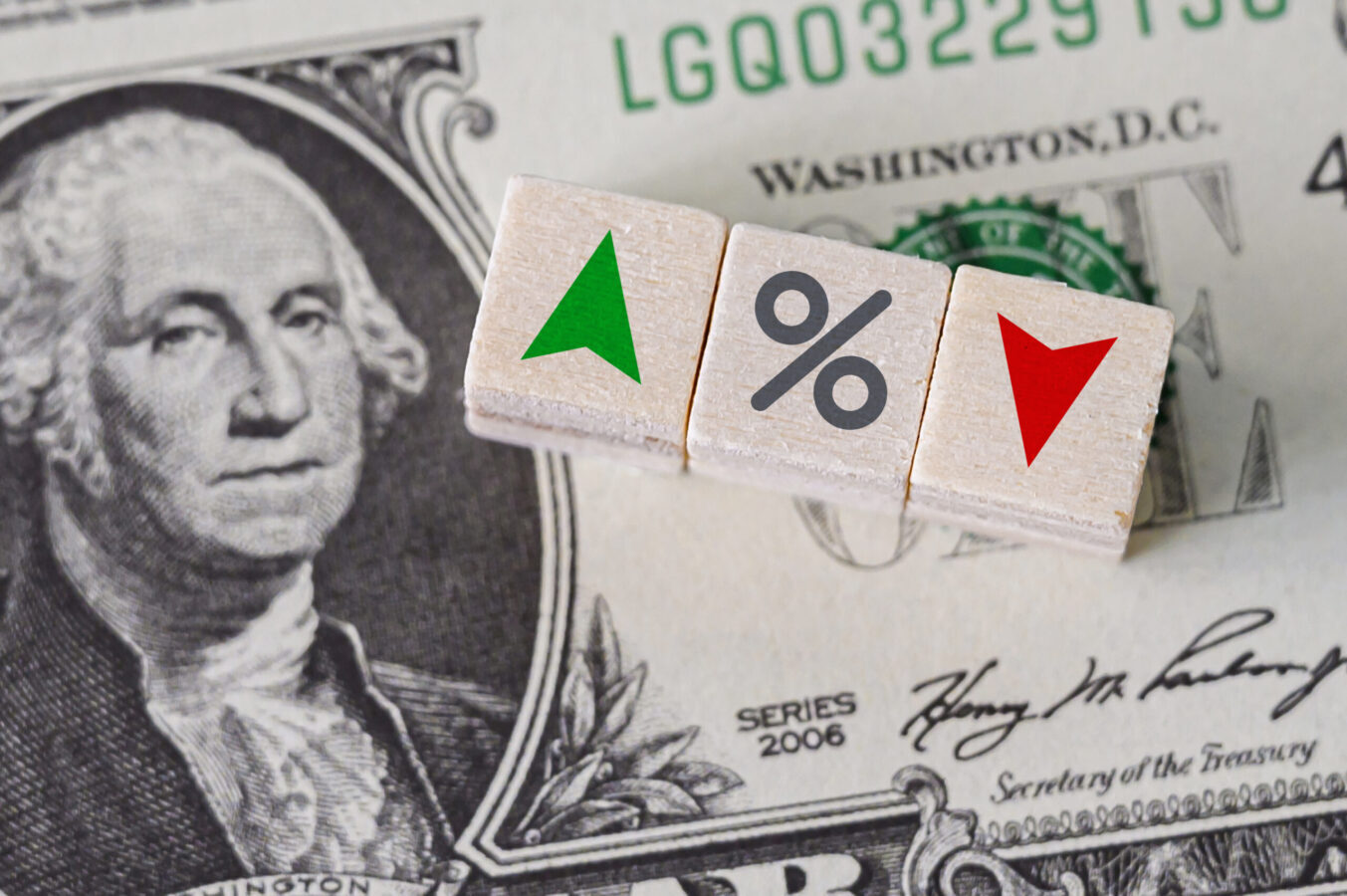 Why Are Stocks Down Today. a photo of a dollar bill with three pieces of dice on it, which show a green "up" arrow, a percentage sign, and a red "down" arrow, respectively