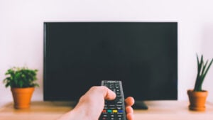 A remote being held and pointed at a black flatscreen tv with two potted plants on either side