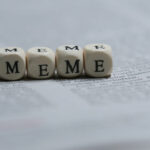 Four dice on a newspaper with letters instead of dots, spelling out the word "Meme". Meme Stocks to Sell