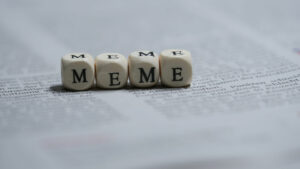 Four dice on a newspaper with letters instead of dots, spelling out the word "Meme"