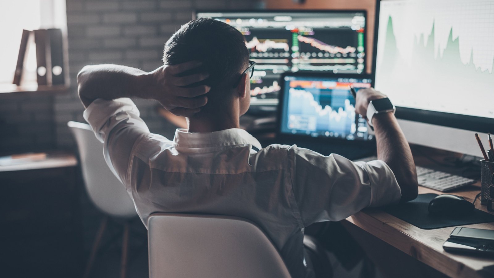 Businessman looking at stock charts on computer screen with one hand on the back of his head and the other hand holding a pen